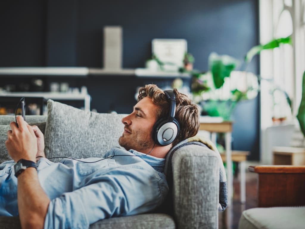 En man ligger avslappnat i soffan med hörlurar på och lyssnar på musik via sin mobiltelefon.