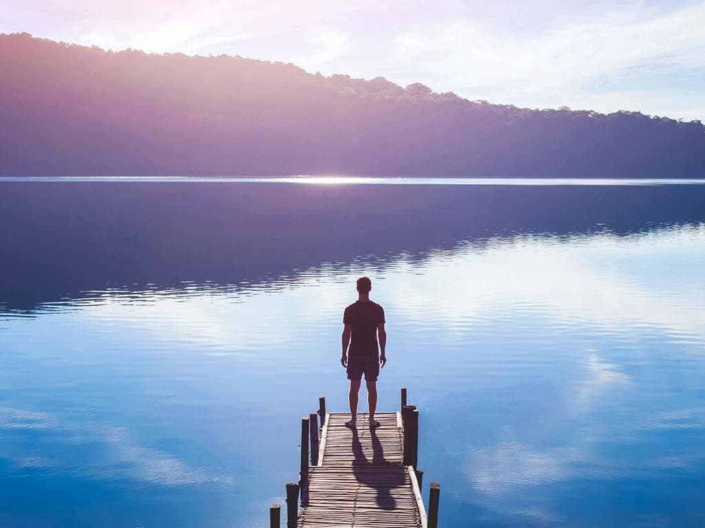  En person står ensam på en träbrygga vid en spegelblank sjö, omgiven av berg i solnedgång.