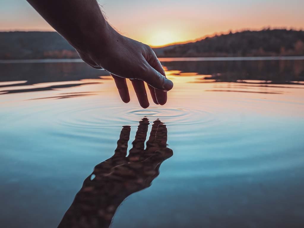 En hand sträcks ner mot en spegelblank sjö vid solnedgång, och reflekteras i vattnet.