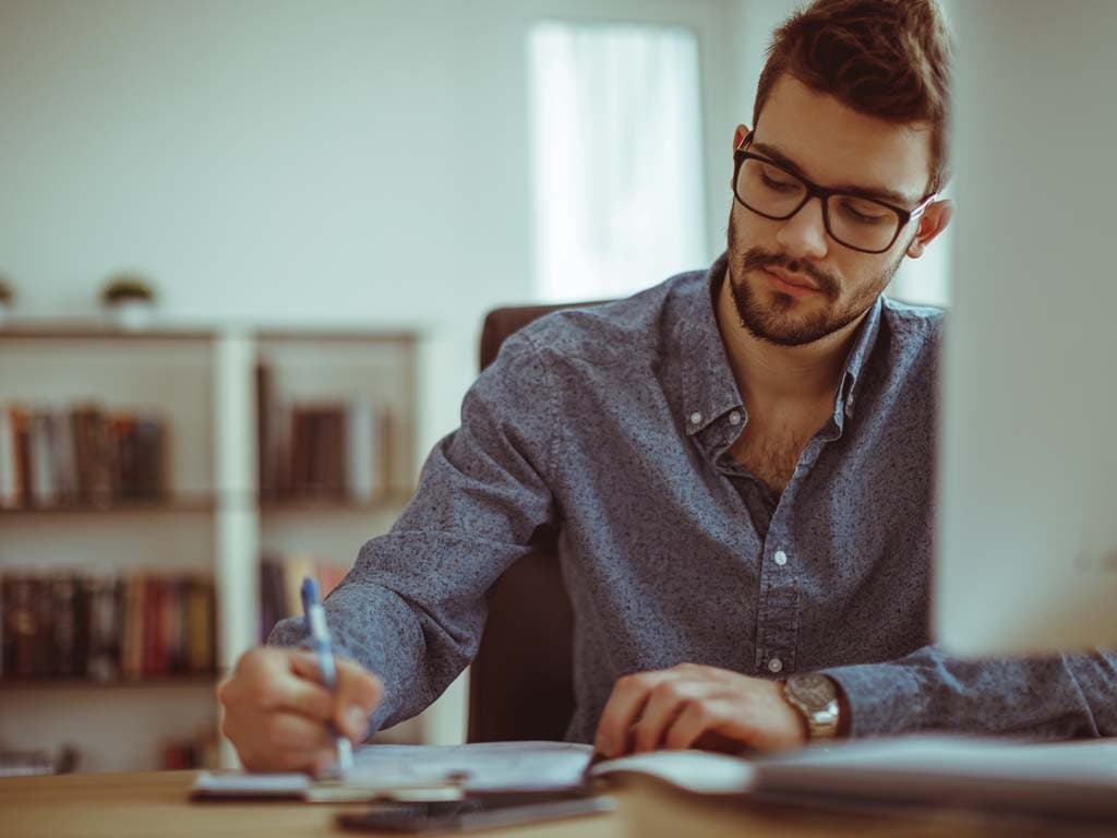  En man sitter vid ett skrivbord hemma och skriver i en anteckningsbok, med laptop och kaffekopp bredvid sig.