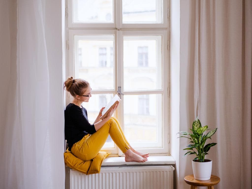 En student sitter i ett fönster med benen uppdragna och läser en bok, omgiven av dagsljus.